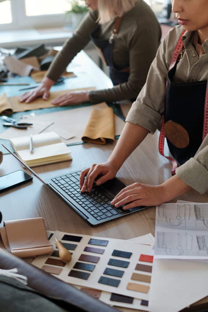 Designer de maroquinerie tapant sur un laptop. Table avec échantillons de cuir, croquis de sacs et portefeuille. Une femme travaille le cuir en arrière-plan.