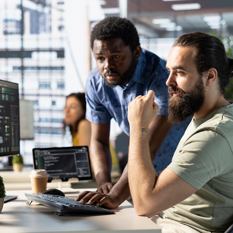 Deux développeurs concentrés sur un écran, un homme noir debout et un homme barbu assis collaborant sur du code informatique en bureau.